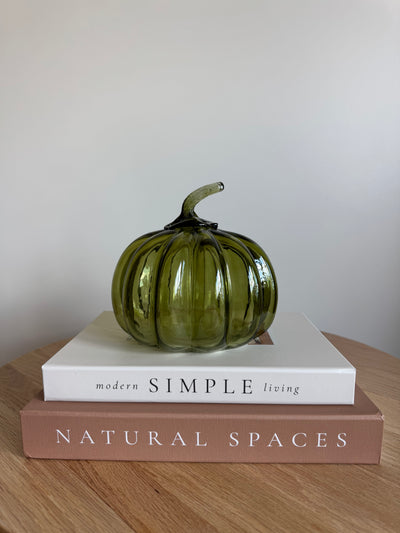 A ribbed, translucent olive green glass pumpkin with a curved stem sits on top of two stacked books on a wooden table against a plain light gray wall.