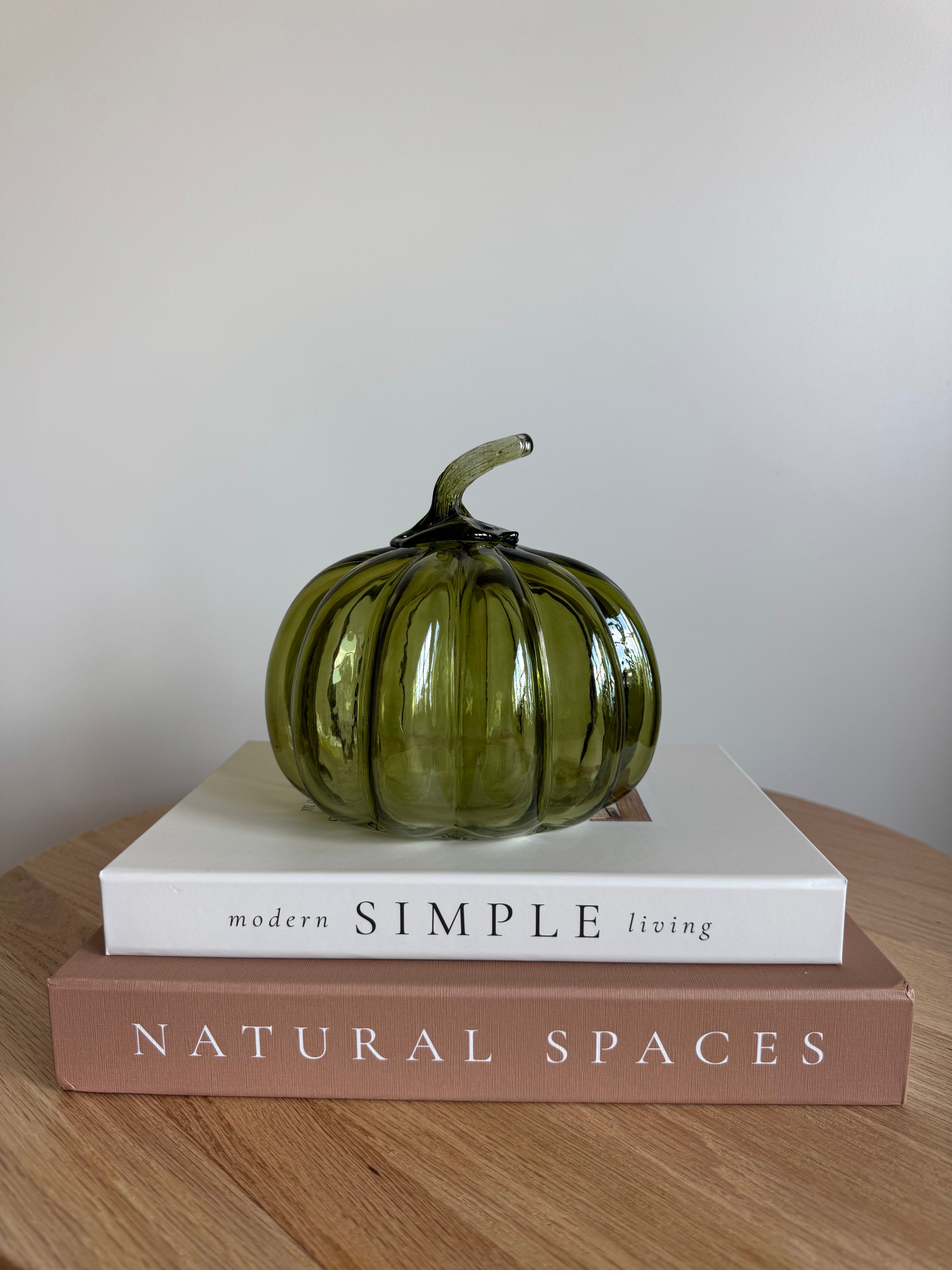 A ribbed, translucent olive green glass pumpkin with a curved stem sits on top of two stacked books on a wooden table against a plain light gray wall.