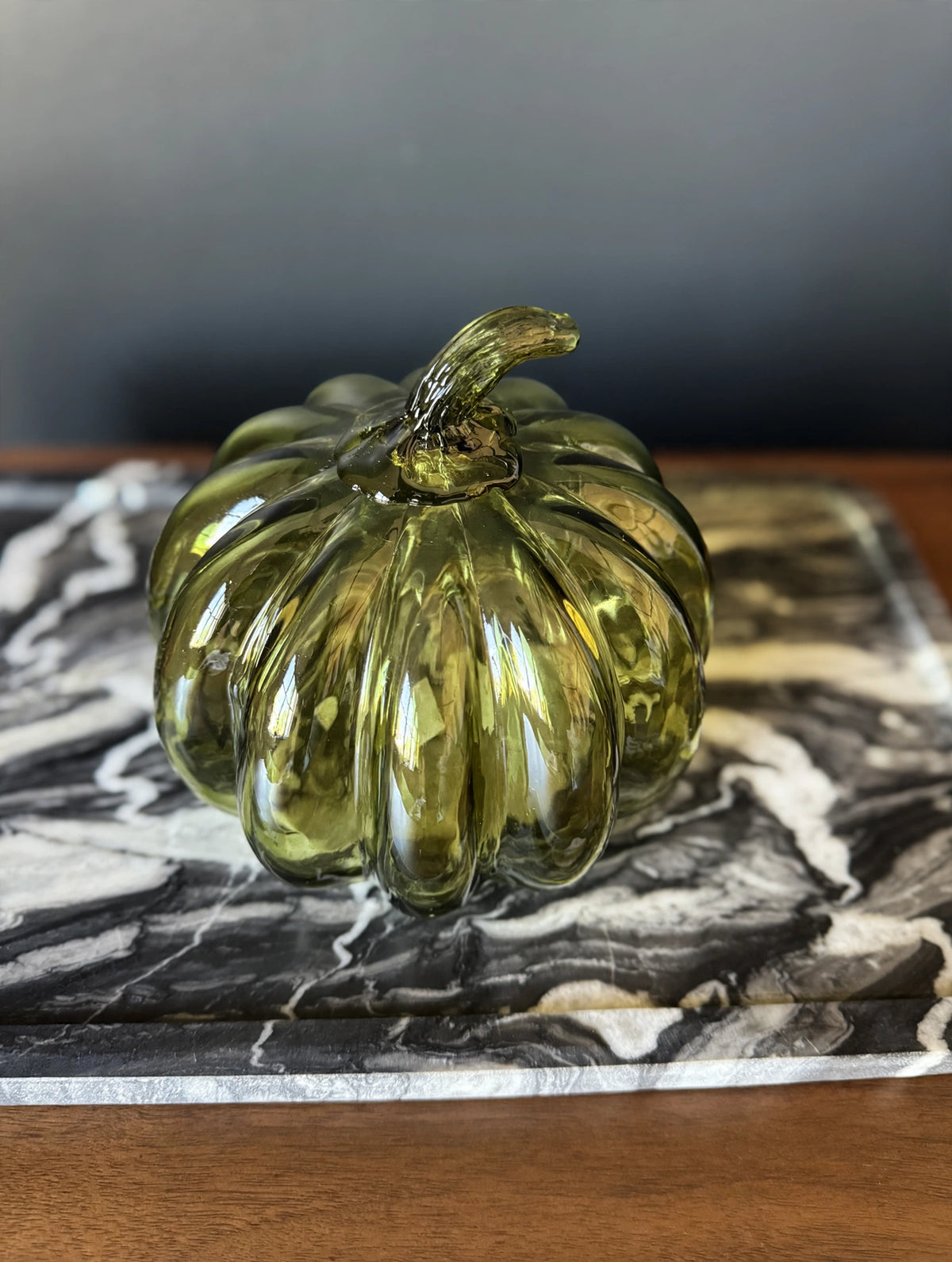 A ribbed, glossy olive green glass pumpkin figurine displayed on a black and white marble tray over a wooden surface.