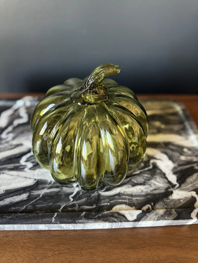 A ribbed, glossy olive green glass pumpkin figurine displayed on a black and white marble tray over a wooden surface.