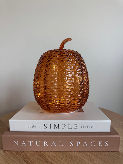An amber glass decorative pumpkin with a textured basketweave pattern sits on top of two stacked books on a wooden surface against a plain light gray wall.